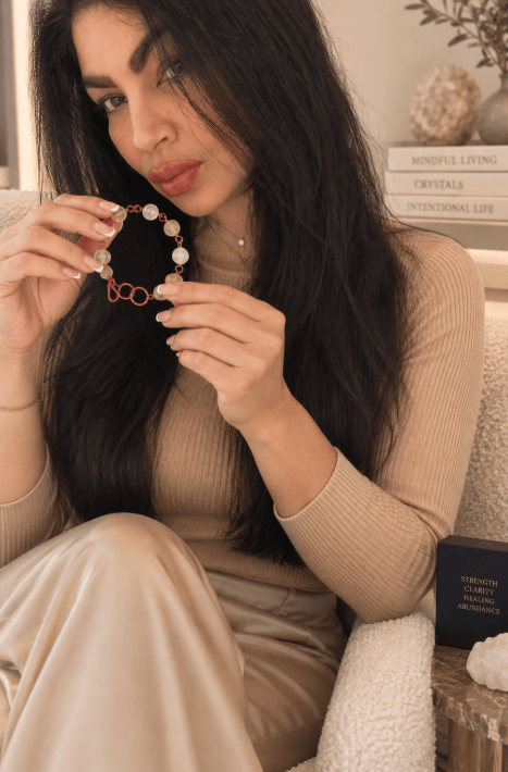 A person holding a bracelet made of beads and links, sitting on a cozy chair with a neutral-coloured outfit. The background includes decorative items and natural light.