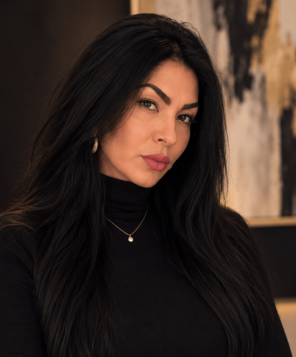 A woman with long black hair and bold eyebrows, wearing a black turtleneck and a pearl necklace, poses confidently against an abstract background.